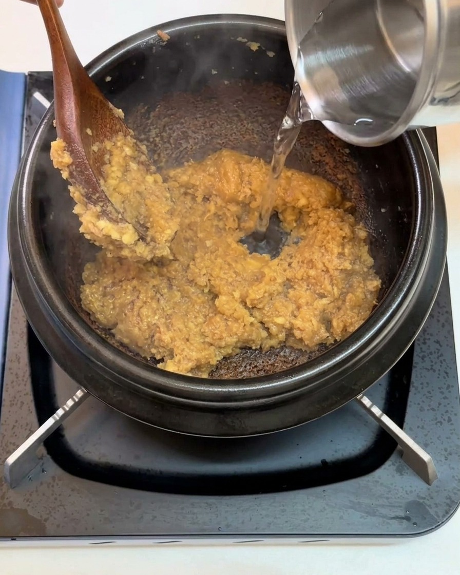 Acqua versata da un contenitore metallico in un tegame nero con la base di pasta di soia soffritta.