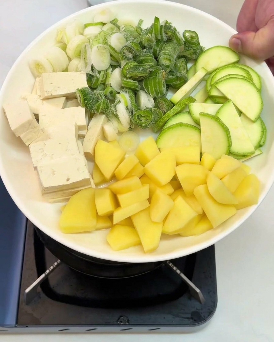 Un piatto bianco con sezioni ben organizzate di tofu a cubetti, cipollotti tritati, zucchine a fette e patate crude a dadini, pronto per essere versato.