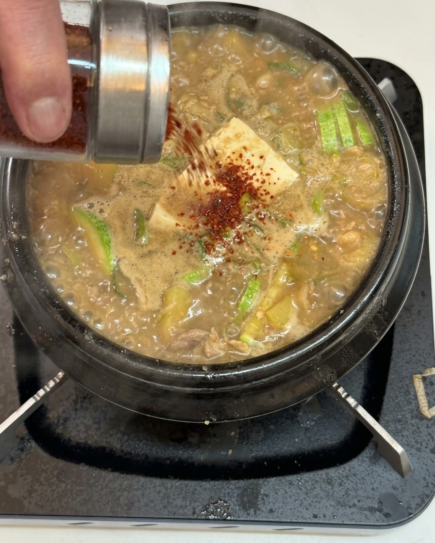 Peperoncino rosso in polvere spolverato da un dosatore di vetro su un bollente stufato coreano con tofu e zucchine.