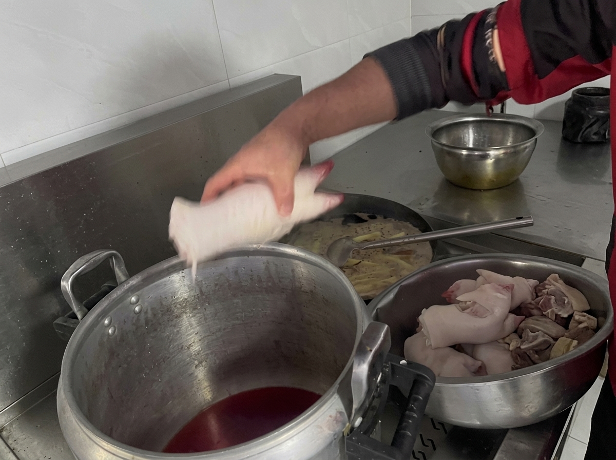 La mano di una persona inserisce gli zampetti di maiale crudi in una grande pentola di alluminio piena di liquido scuro per brasato.