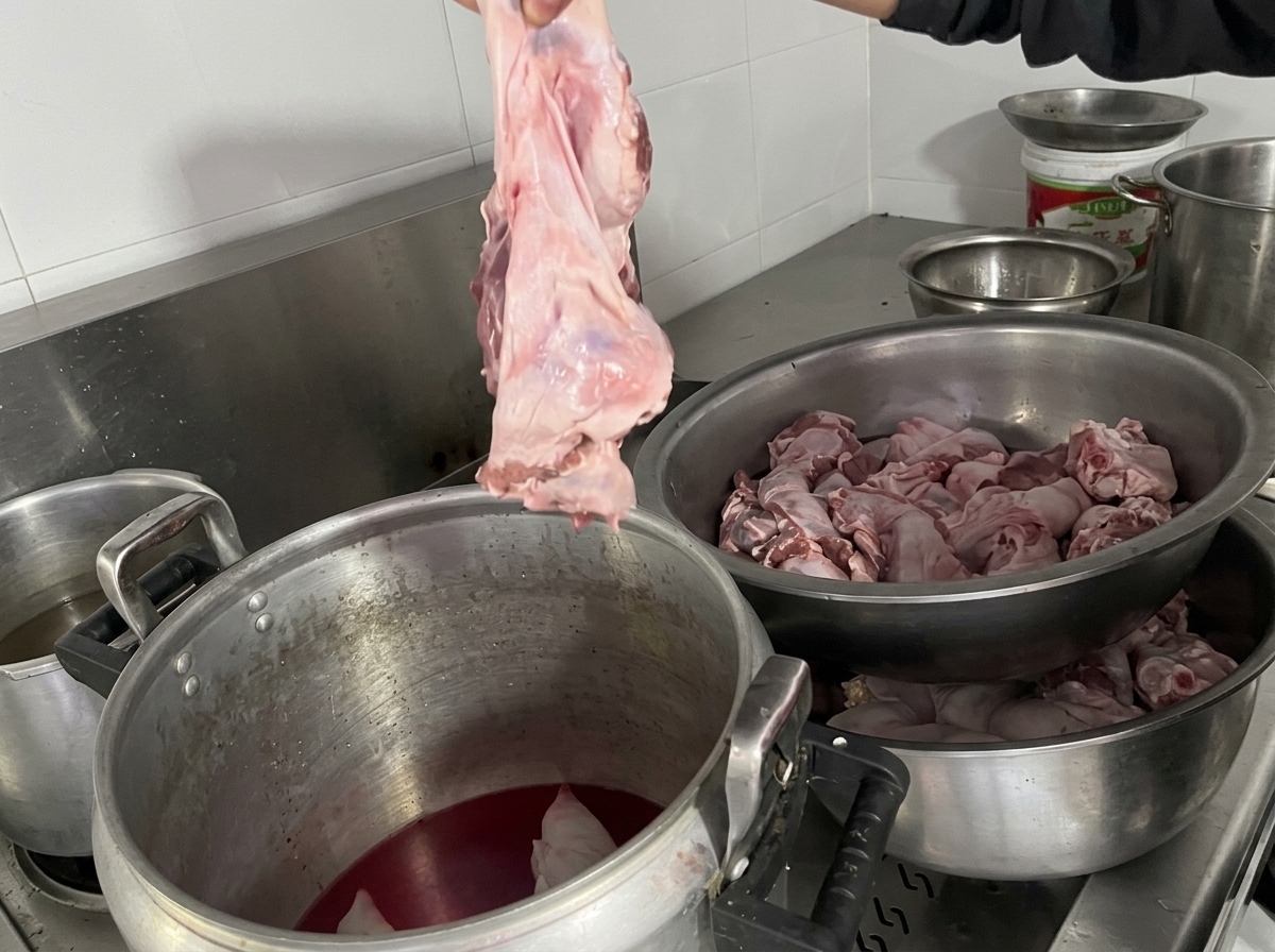 Una mano tiene su un grosso pezzo di diaframma crudo, pronta a immergerlo in una pentola contenente zampetti di maiale e brodo.