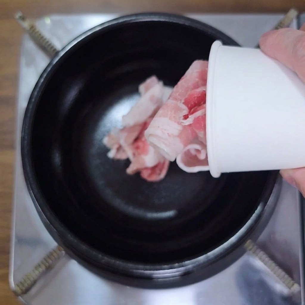 Mano che versa pancetta cruda a fette sottili da un bicchiere di carta in una pentola nera.