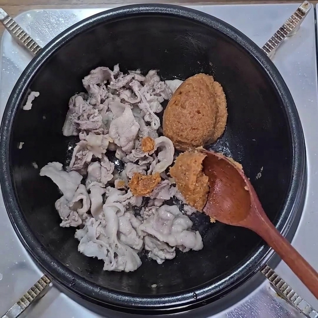 Cucchiai di pasta di soia marrone e ssamjang aggiunti alla pancetta cotta in pentola.