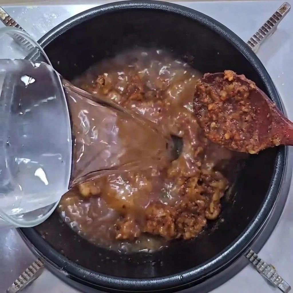 Acqua limpida versata da un contenitore di vetro in una pentola contenente un mix sfrigolante di carne e pasta di soia.
