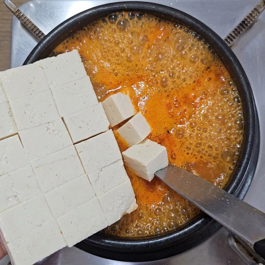 Tofu morbido a cubetti fatto scivolare delicatamente da un grande coltello in una pentola di zuppa bollente.