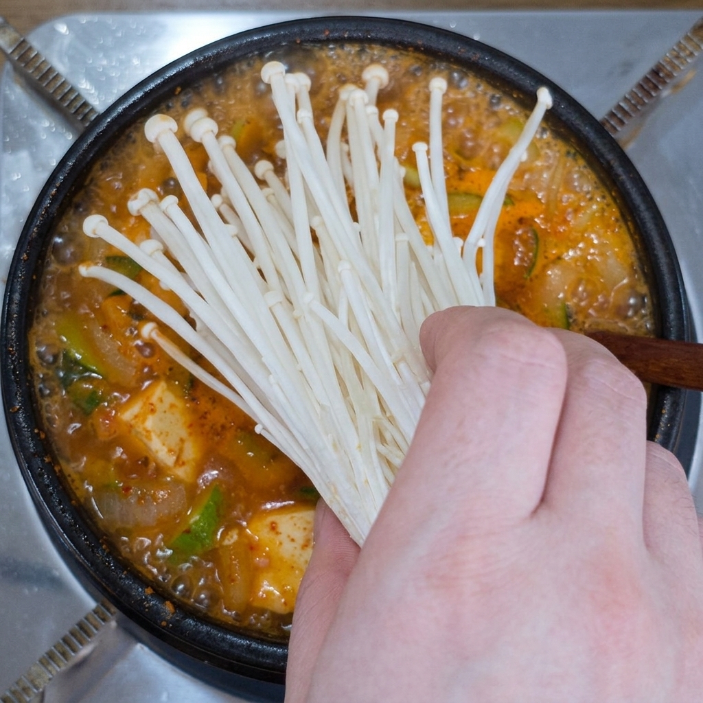 Mano che mette un mazzetto di funghi enoki freschi in una pentola di zuppa di pasta di soia bollente.