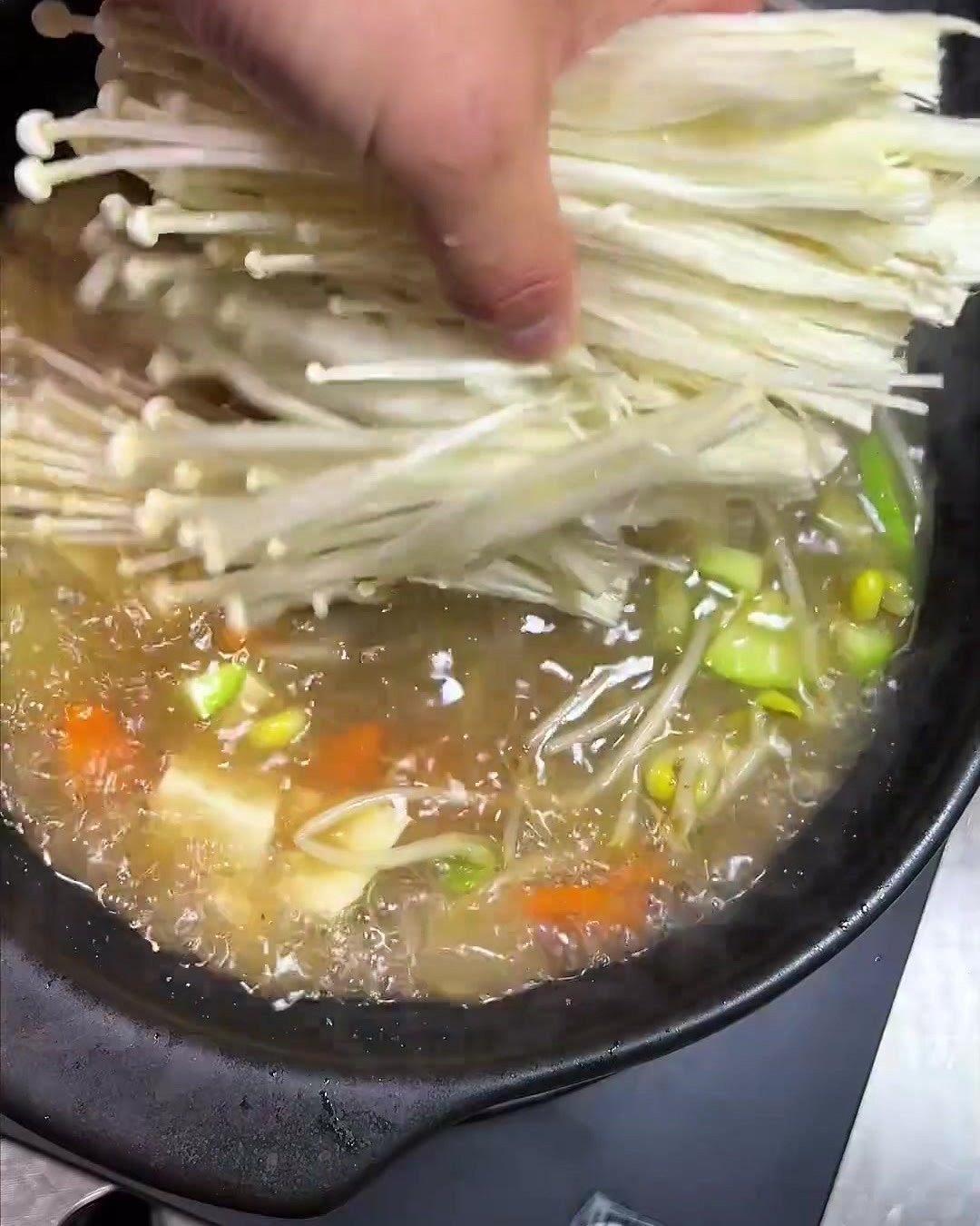 Un mazzetto di delicati funghi enoki a gambo lungo aggiunti alla pentola che sobbolle.