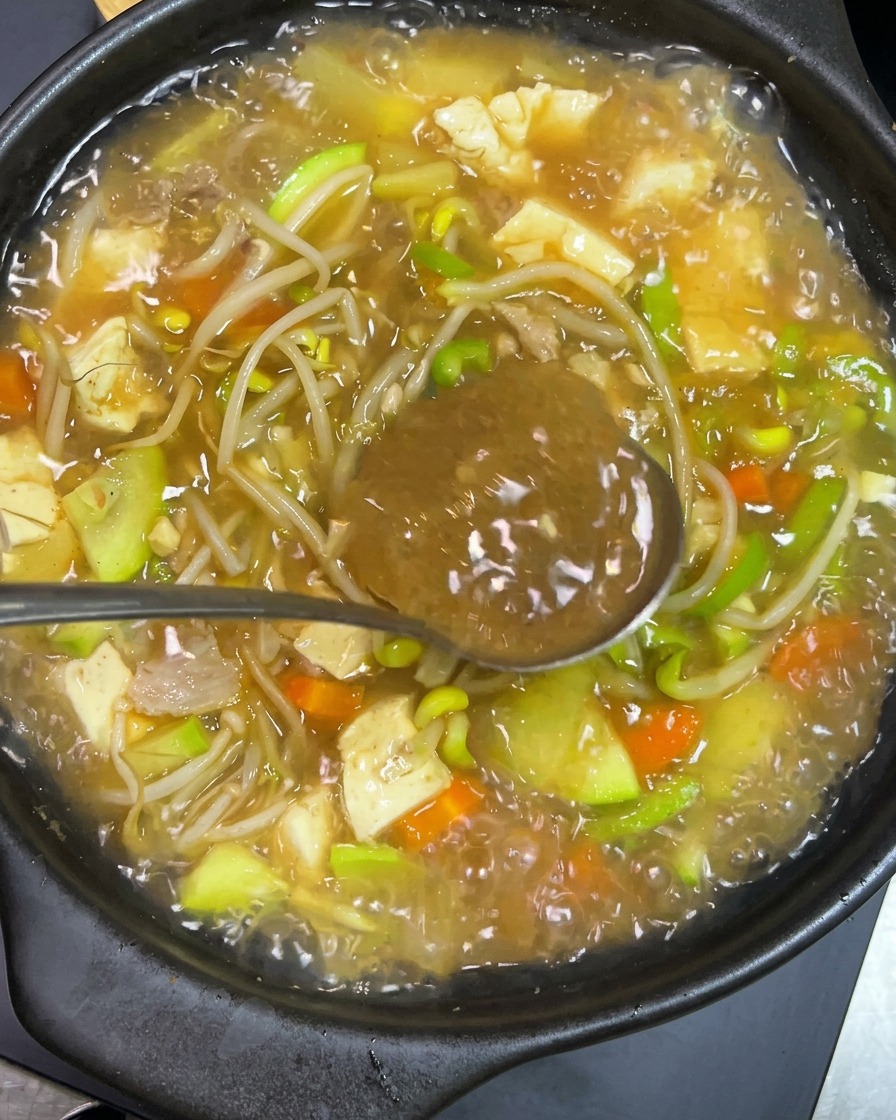 Un cucchiaio di metallo che tiene pasta piccante coreana scura mentre viene mescolata in una pentola di terracotta che gorgoglia riempita di tofu, zucchine, germogli di soia e brodo.