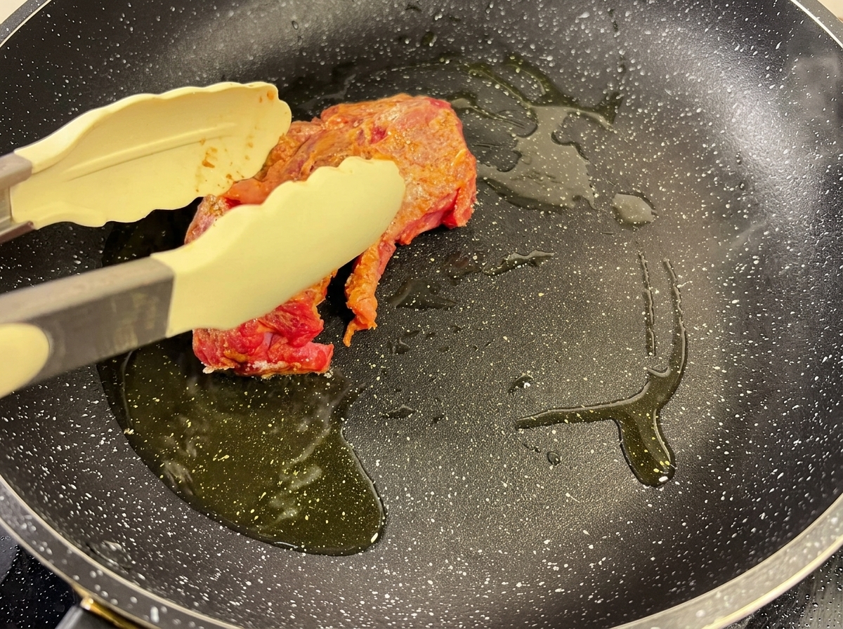 Un pezzo di manzo speziato viene rosolato in una padella calda con olio, tenuto fermo da una pinza.