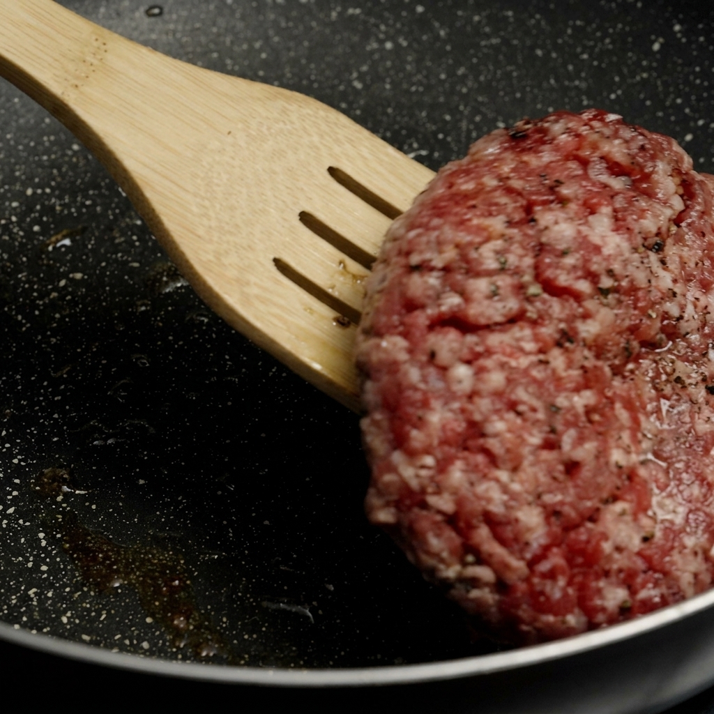 Una spatola di legno solleva e capovolge un medaglione di carne spesso con una bella crosticina bruna in una padella ben calda.