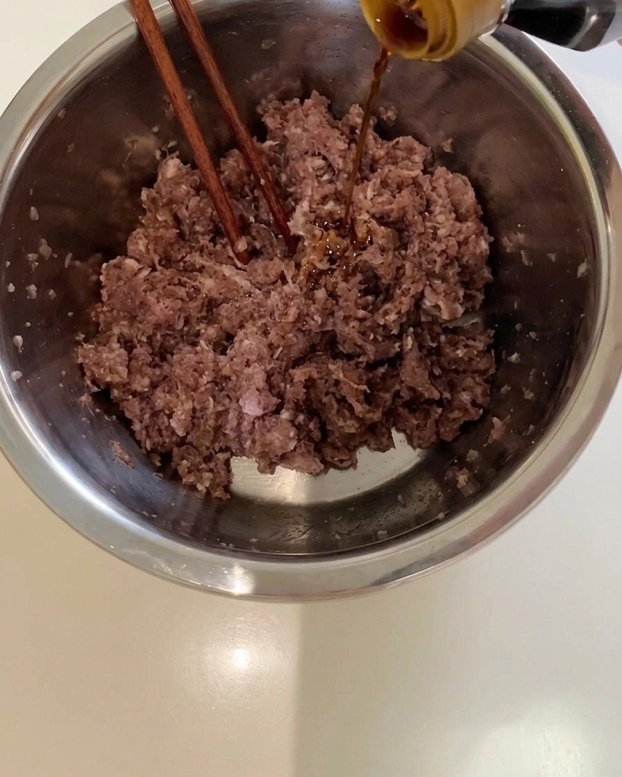 Salsa di soia chiara versata da una bottiglia in una ciotola di metallo piena di carne di manzo macinata.