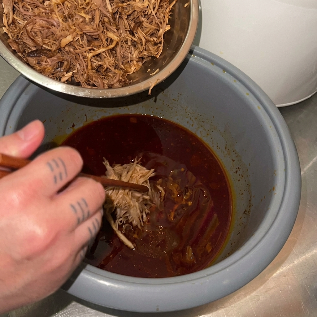Maiale sfilacciato trasferito da una ciotola di metallo di nuovo nel cestello del cuociriso con la salsa rossa scura.