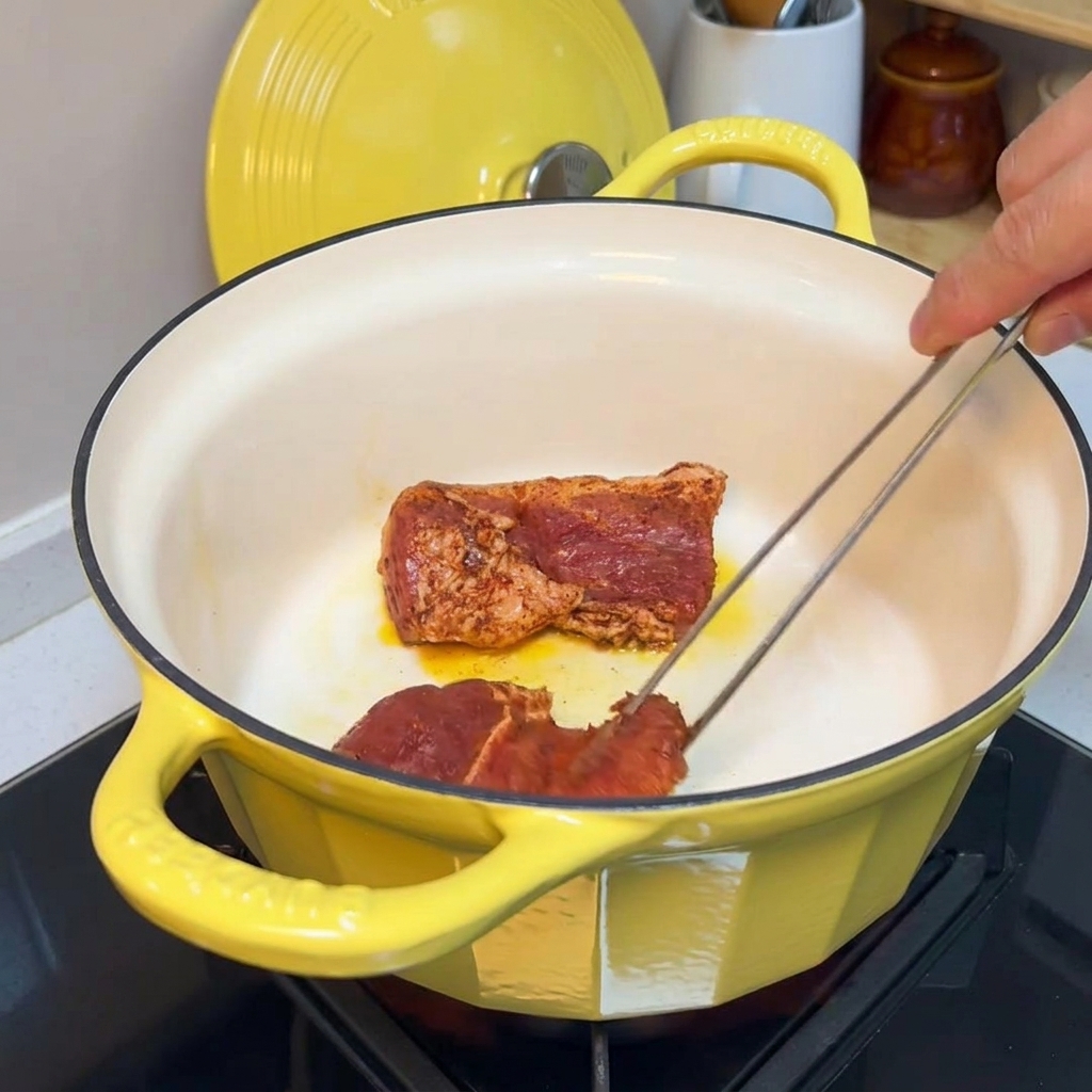 Pinze che girano pezzi di maiale condito in una pentola di ghisa gialla per rosolare i bordi.