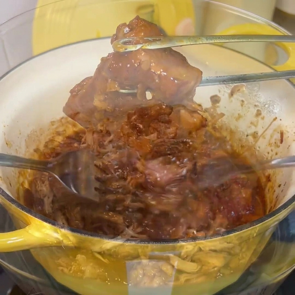 Carne di maiale cotta e tenera sfilacciata con due forchette in una ciotola di vetro.