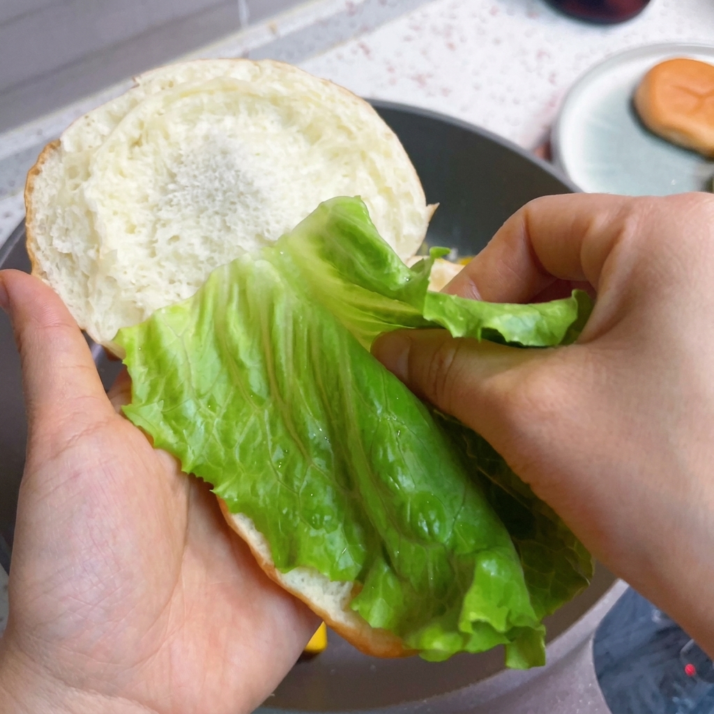 Assemblaggio del burger con lattuga fresca, bacon e uova soffici sul panino.