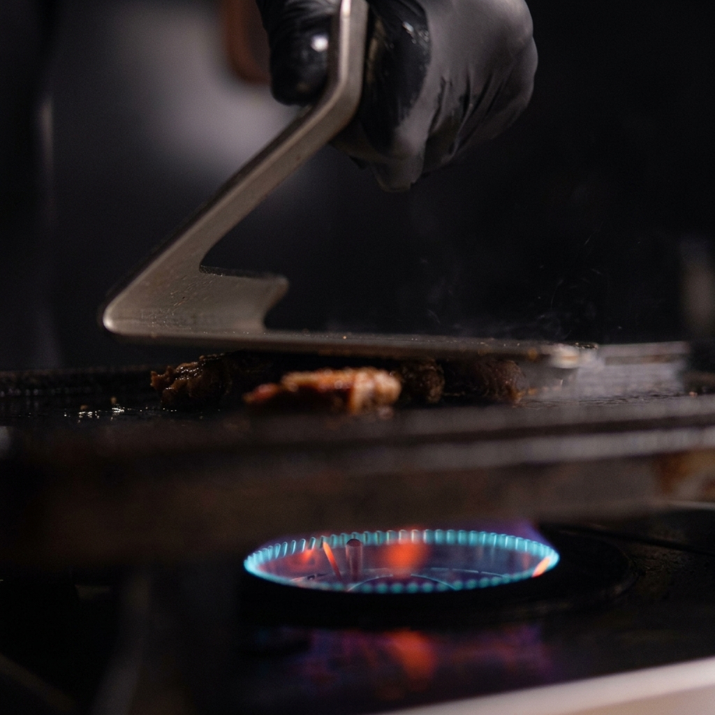 Una mano che utilizza una pressa in metallo per schiacciare un mucchietto di pulled pork su una piastra in ghisa rovente con fiamme blu visibili sotto.