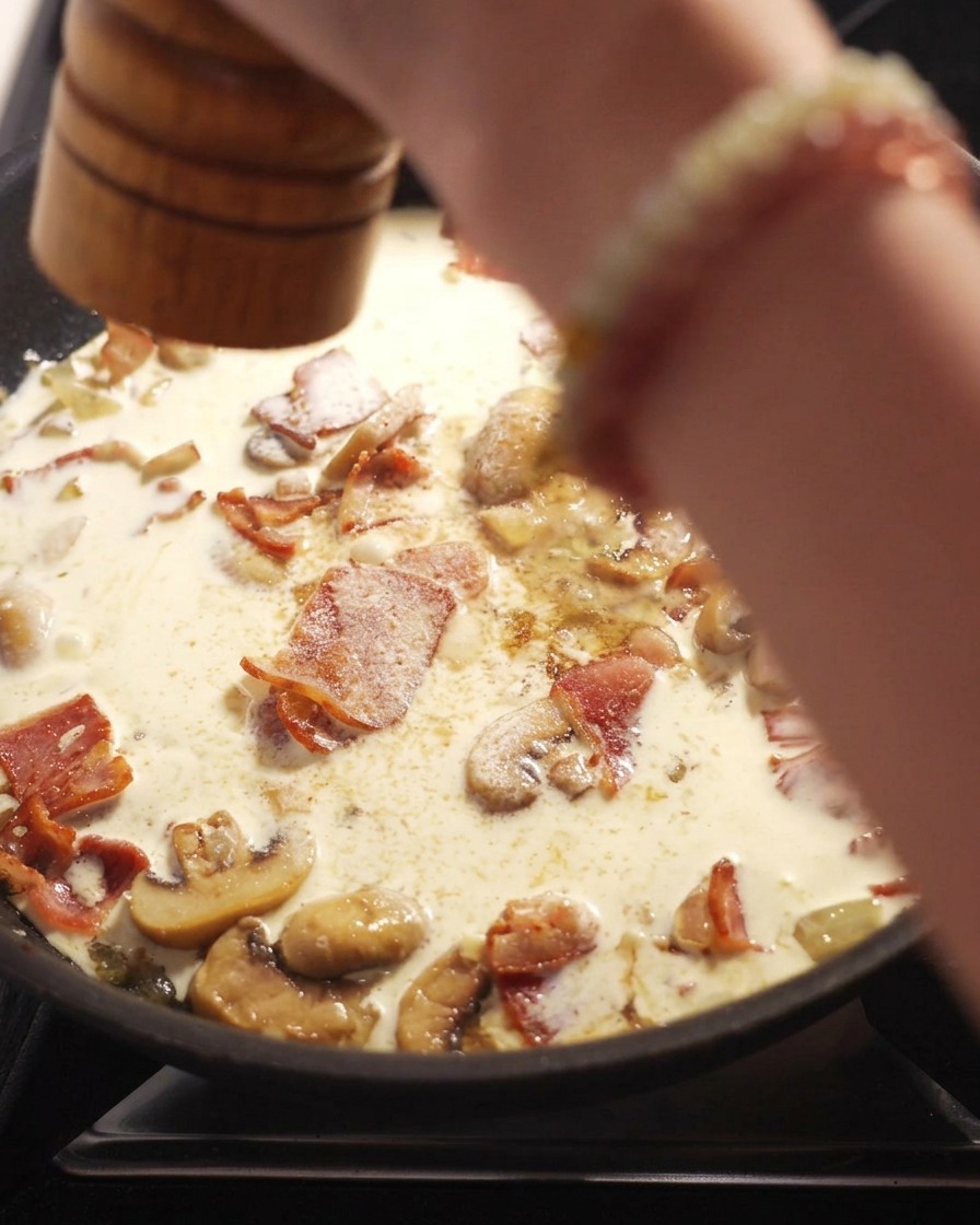 Una mano usa un macinapepe in legno per macinare pepe nero fresco su una padella con salsa cremosa, pancetta e funghi a fette.