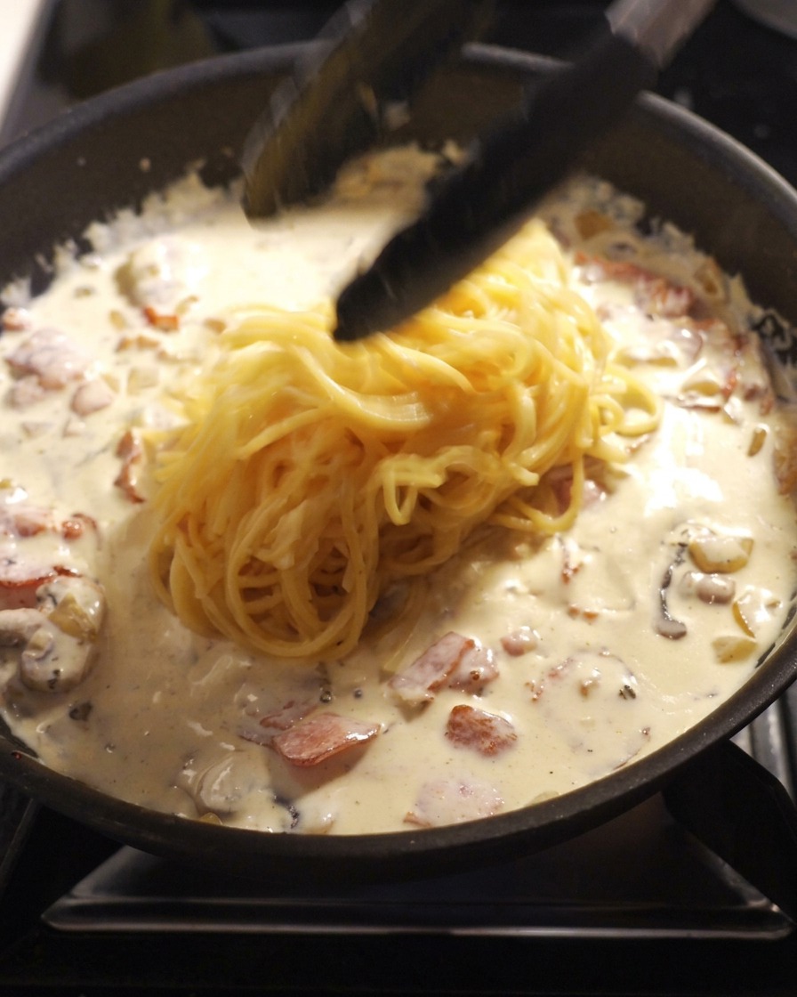 Pinze da cucina nere che tuffano una porzione di spaghetti appena cotti in una padella piena di salsa alla panna, pezzetti di pancetta e funghi.