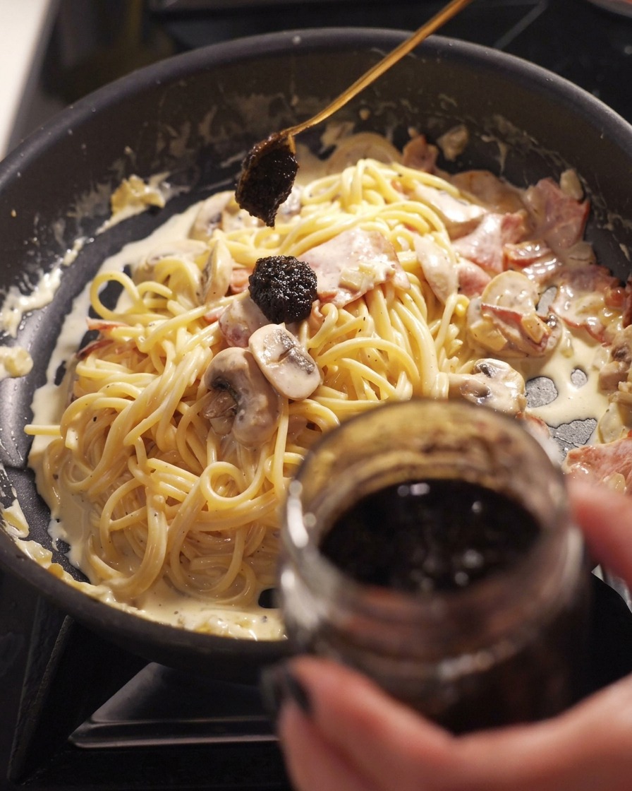 Un cucchiaio fa cadere una scura cucchiaiata di salsa al tartufo nero da un vasetto a una padella di spaghetti cremosi con pancetta e funghi.