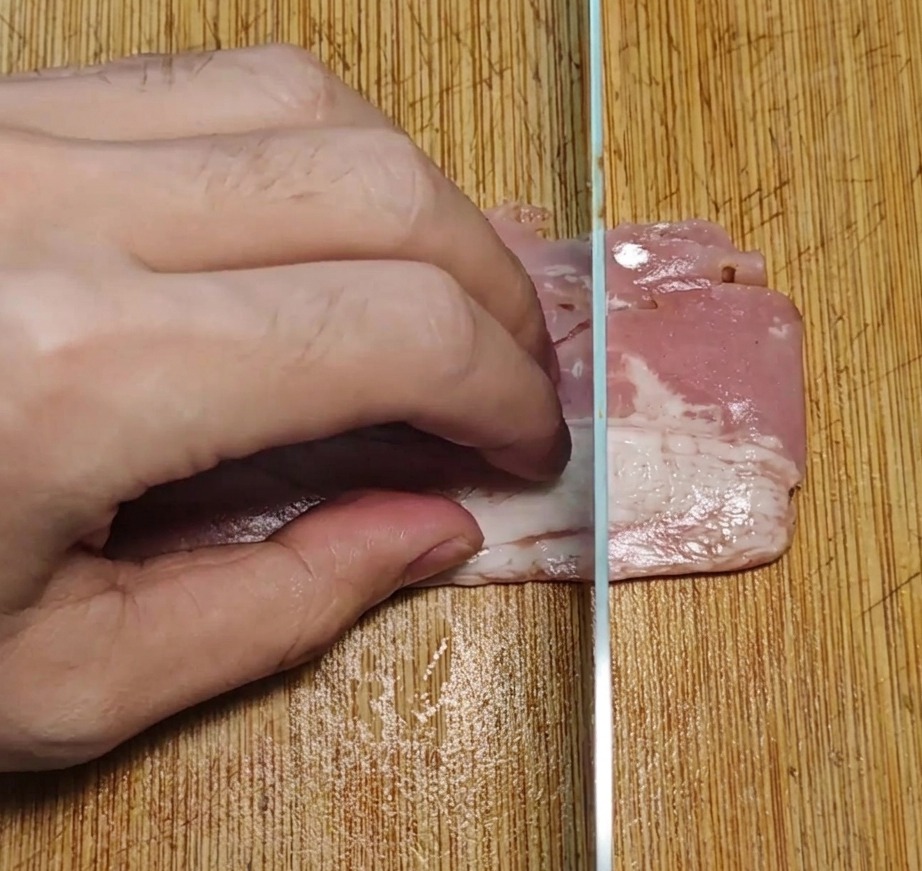 Strisce di pancetta tagliate in pezzi rettangolari uniformi su un tagliere di legno con un coltello da cucina.
