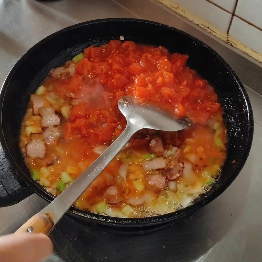 Una spatola schiaccia i pomodori in padella per creare una salsa omogenea.
