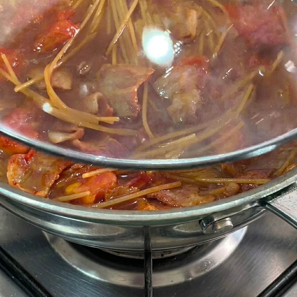 Spaghetti che cuociono nel sugo coperti da un coperchio di vetro.