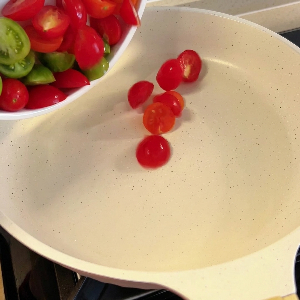 Pomodorini tagliati a metà aggiunti in una padella bianca.