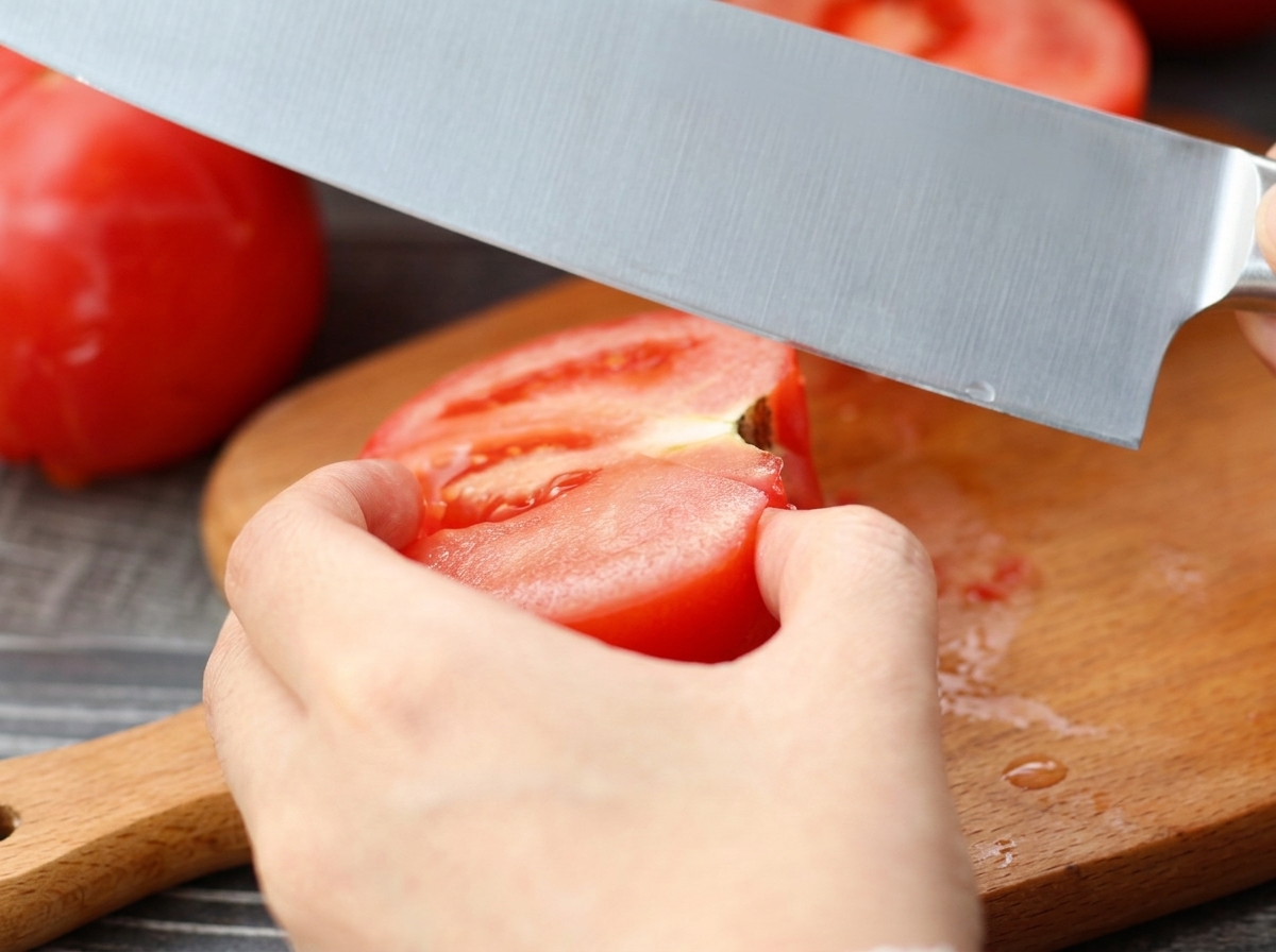 Un coltello che taglia un pomodoro sbucciato su un tagliere di legno.