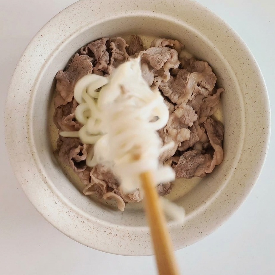 Ciotola con fettine di manzo alla base, sormontate da udon freddi e funghi enoki.