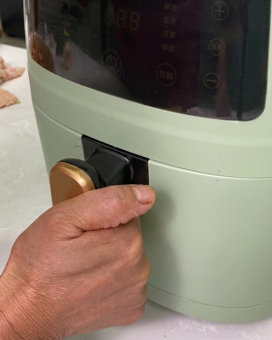 Una mano che inserisce una vaschetta di alluminio piena di involtini speziati in una friggitrice ad aria verde.