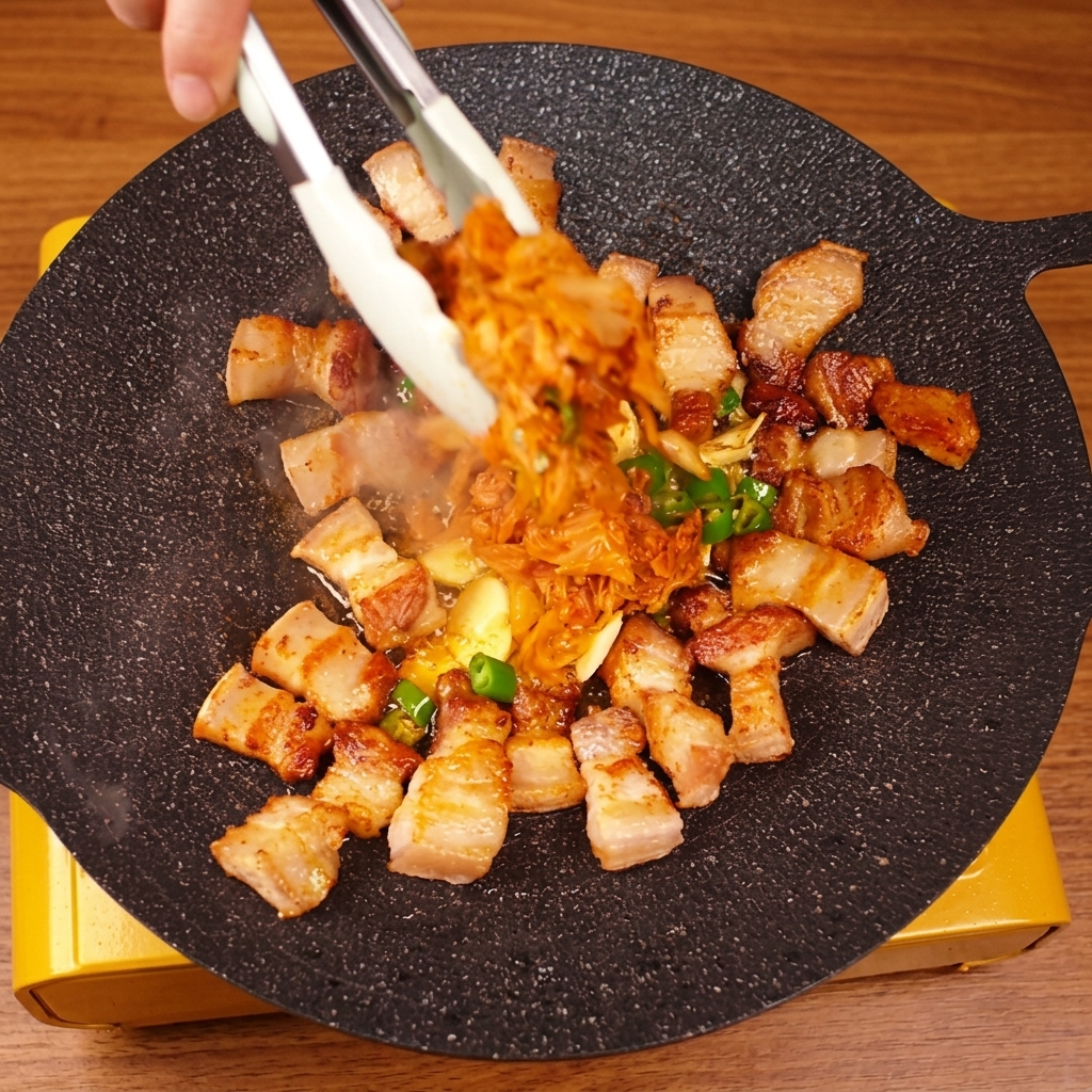 Una mano usa le pinze per mescolare il maiale e il kimchi sulla piastra calda.