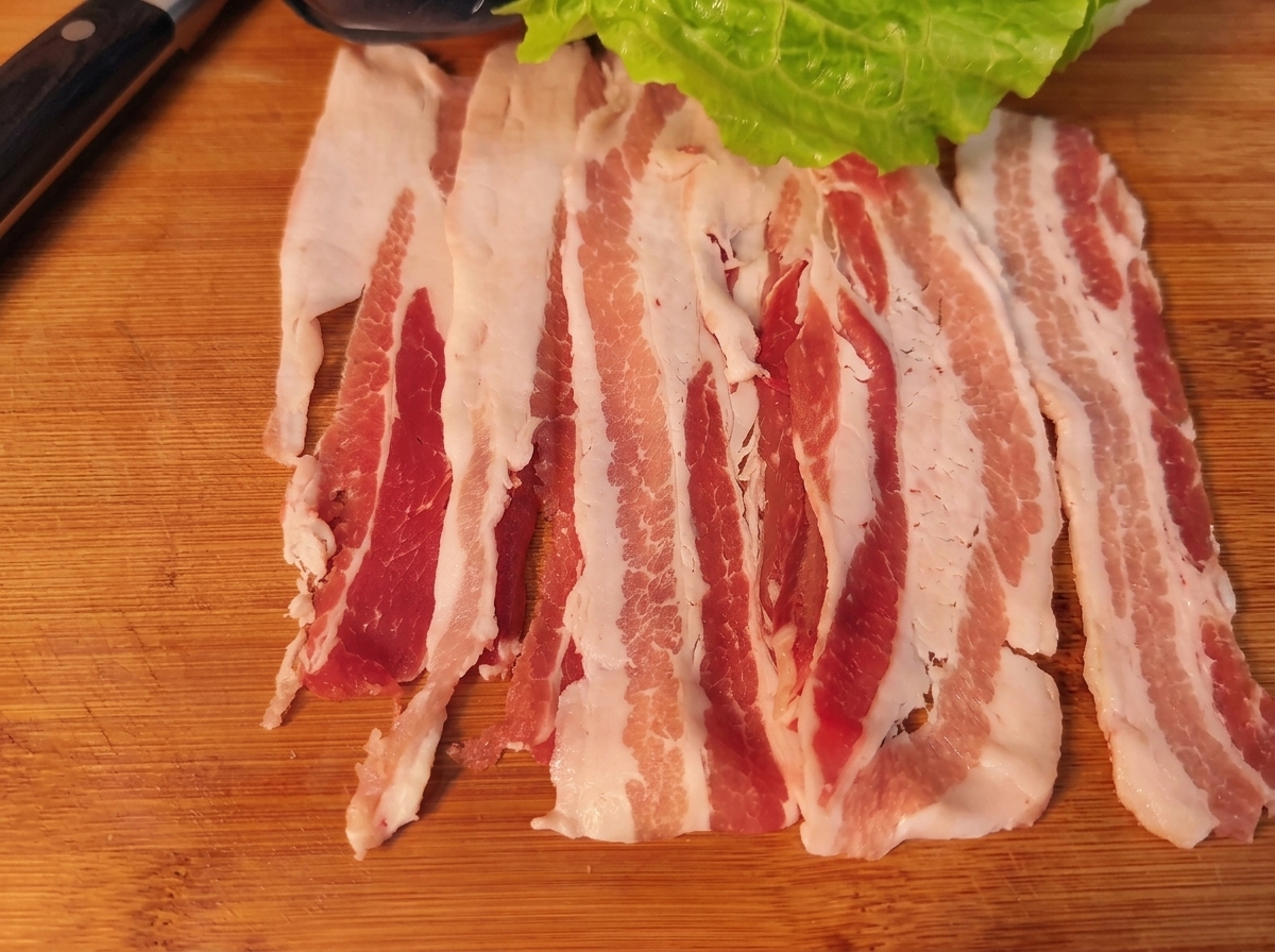 Una fetta di pancetta guarnita con una foglia di lattuga, carota, cipolla e kimchi, pronta per essere arrotolata.