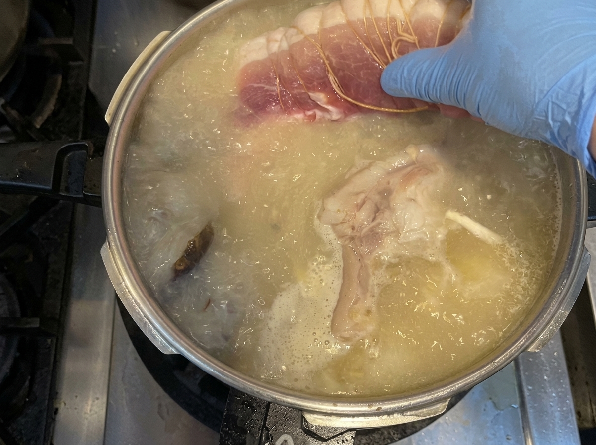 Mano con guanto blu che inserisce la spalla di maiale legata nel brodo con le verdure.