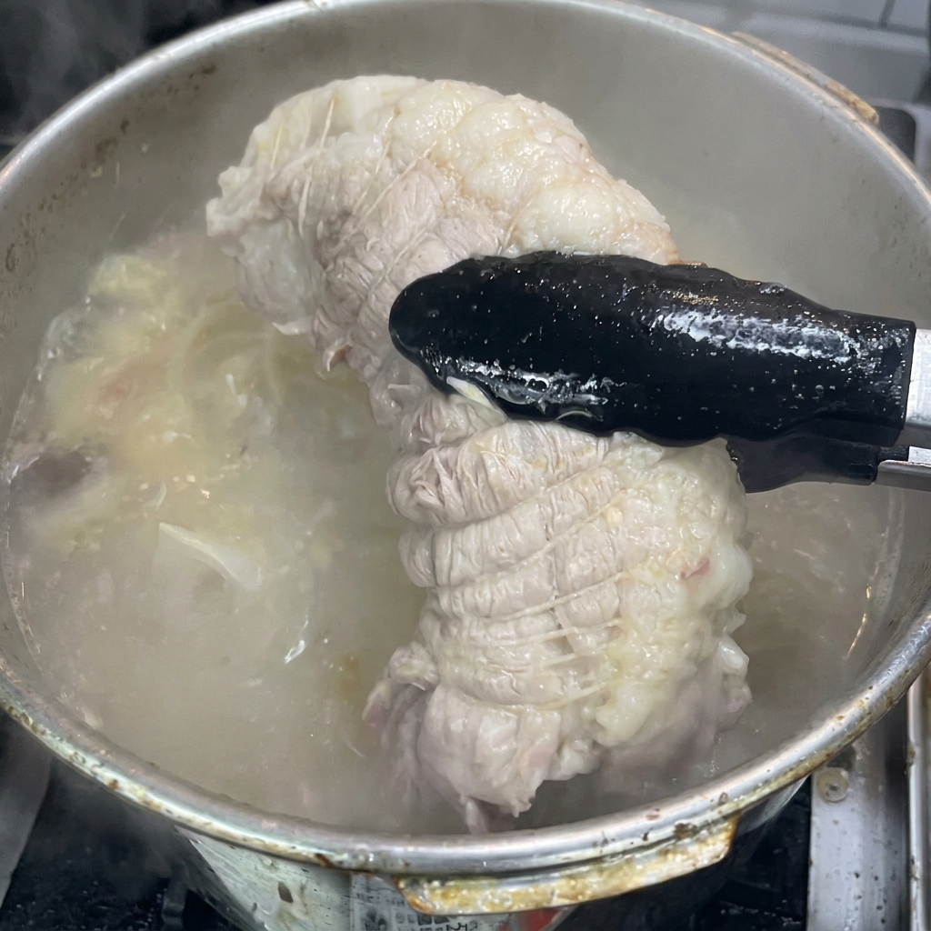 Pinze nere che sollevano la spalla di maiale cotta dal brodo bollente.