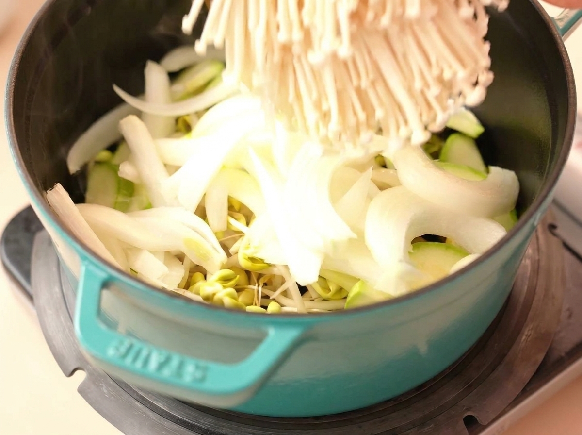 Un mazzetto di funghi enoki crudi inserito in una pentola con cipolla, zucchine e germogli.