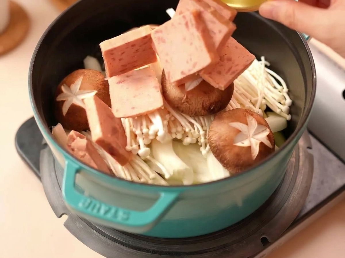 Cubetti di Spam aggiunti in una pentola con funghi enoki, cipolle e funghi shiitake incisi.