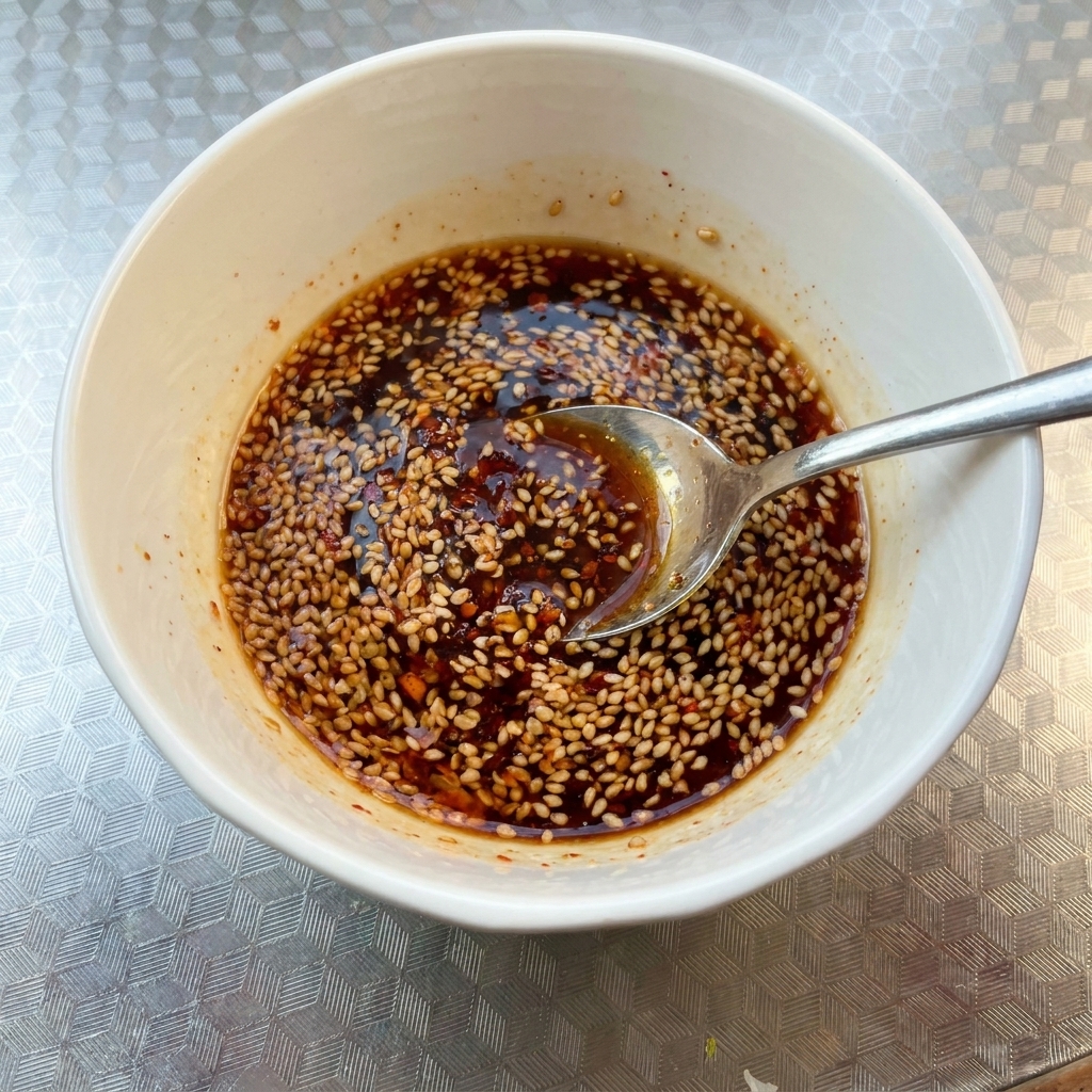 Una ciotola bianca contenente una miscela di salsa scura e speziata guarnita con semi di sesamo, mescolata con un cucchiaio.