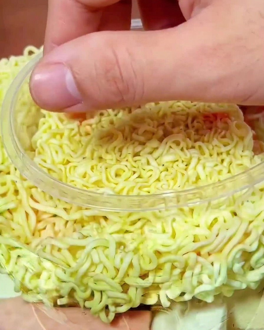 Una mano che mette pezzi di kimchi rosso vibrante sopra i noodles crudi nella pentola.