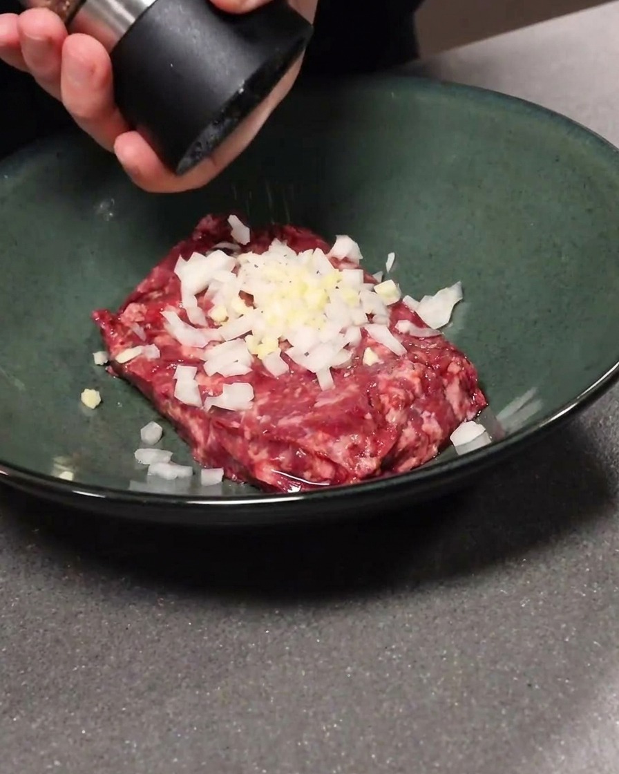 Carne macinata mescolata con aglio e cipolla in una ciotola, mentre viene aggiunto pepe nero.