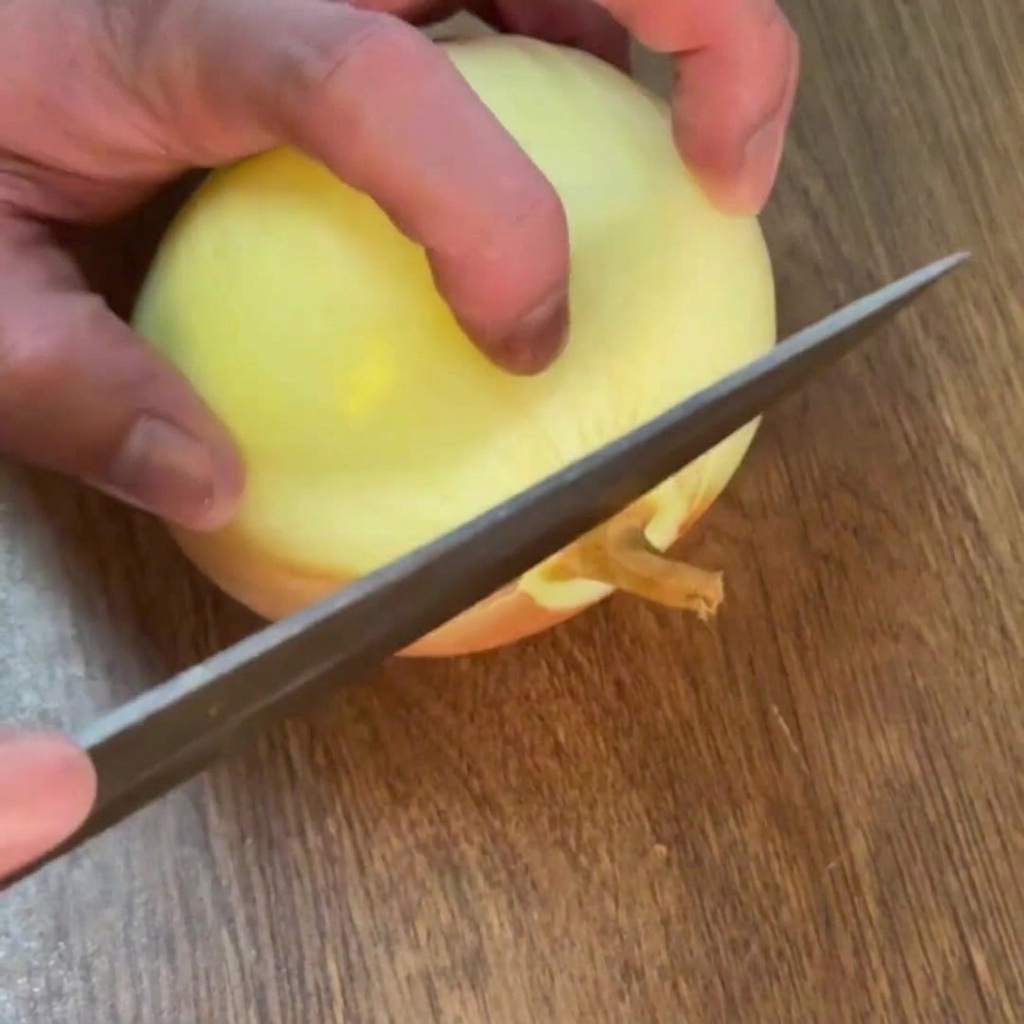 Mani che usano un coltello per rimuovere la radice di una cipolla dorata sbucciata su un tagliere in legno.
