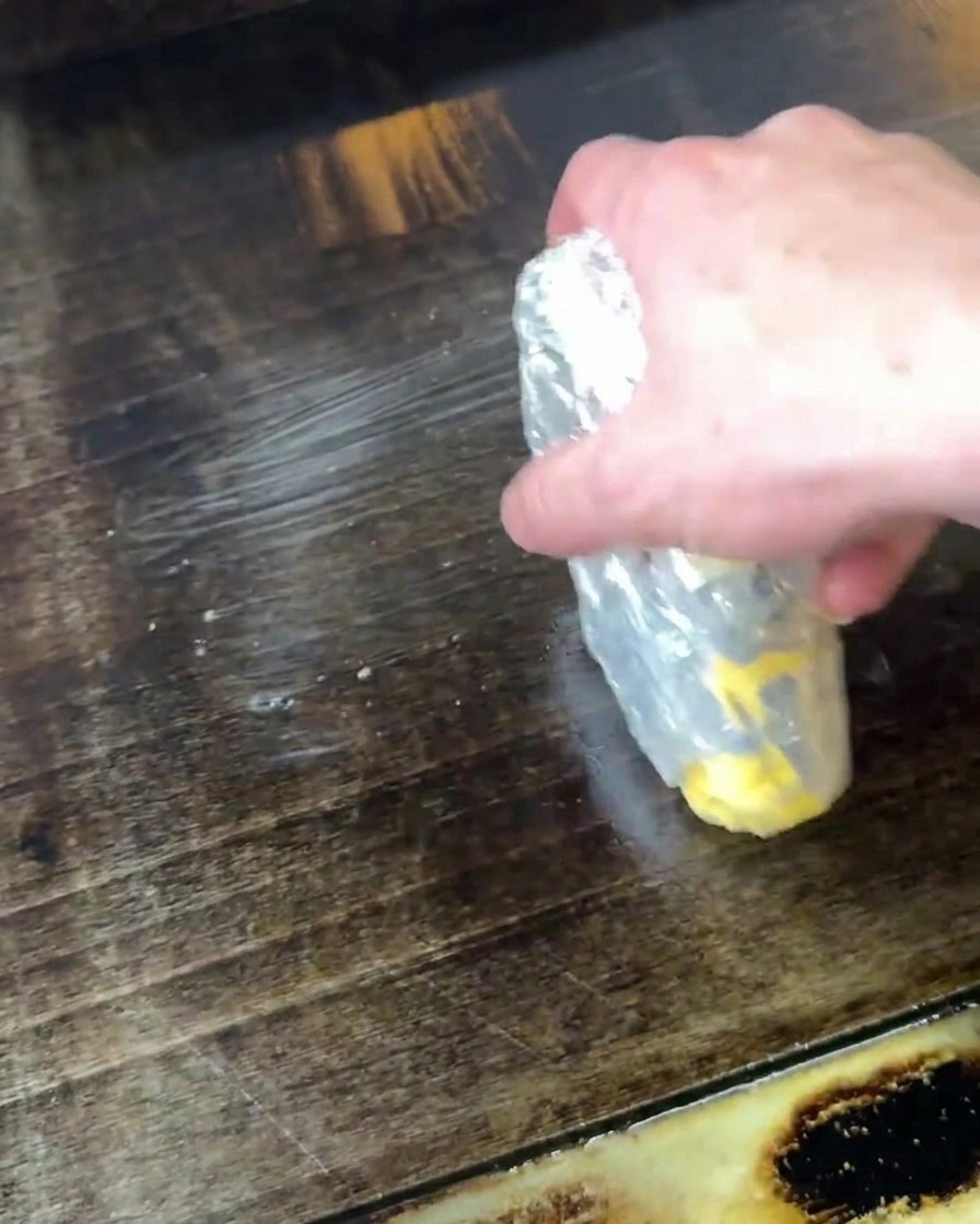 Una mano che tiene un panetto di burro parzialmente scartato, sciogliendolo strofinandolo direttamente su una piastra calda scura.