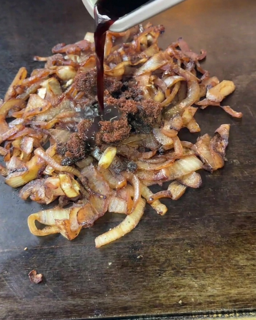 Aceto invecchiato scuro e grumi di zucchero di canna versati da un contenitore di metallo su cipolle in cottura.