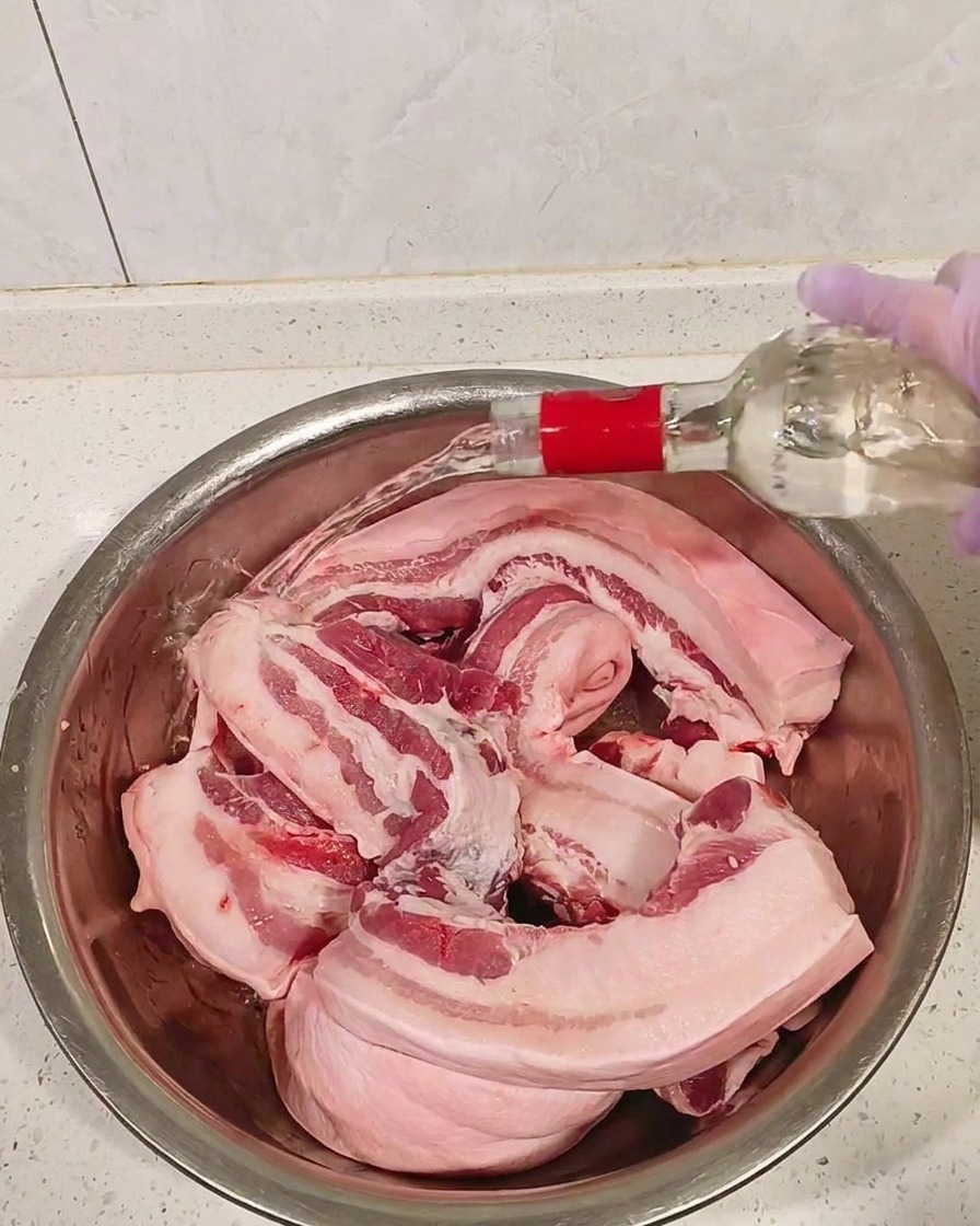 Pezzi di pancetta cruda in una ciotola di metallo bagnati con distillato trasparente.