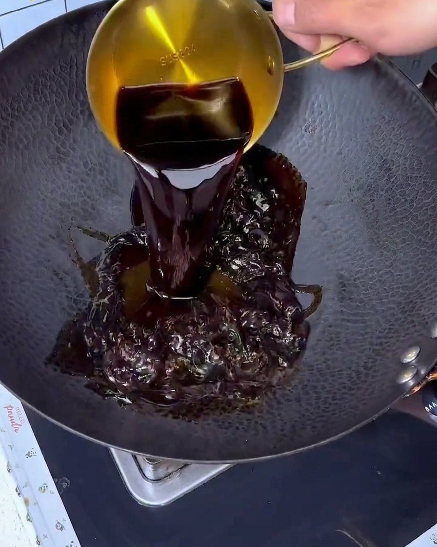 Salsa di soia versata da un misurino dorato in un grande wok nero.