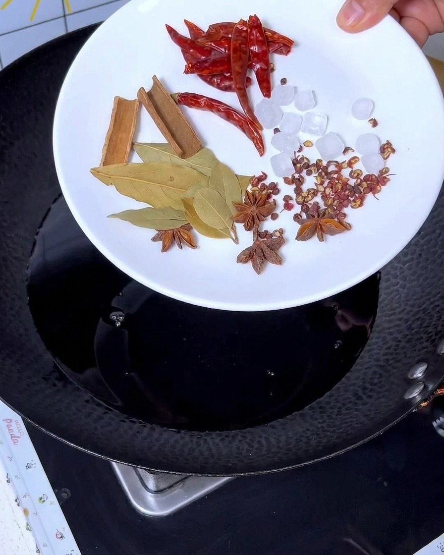 Spezie come cannella, anice stellato, alloro, peperoncino e zucchero grezzo aggiunte al liquido scuro nel wok.