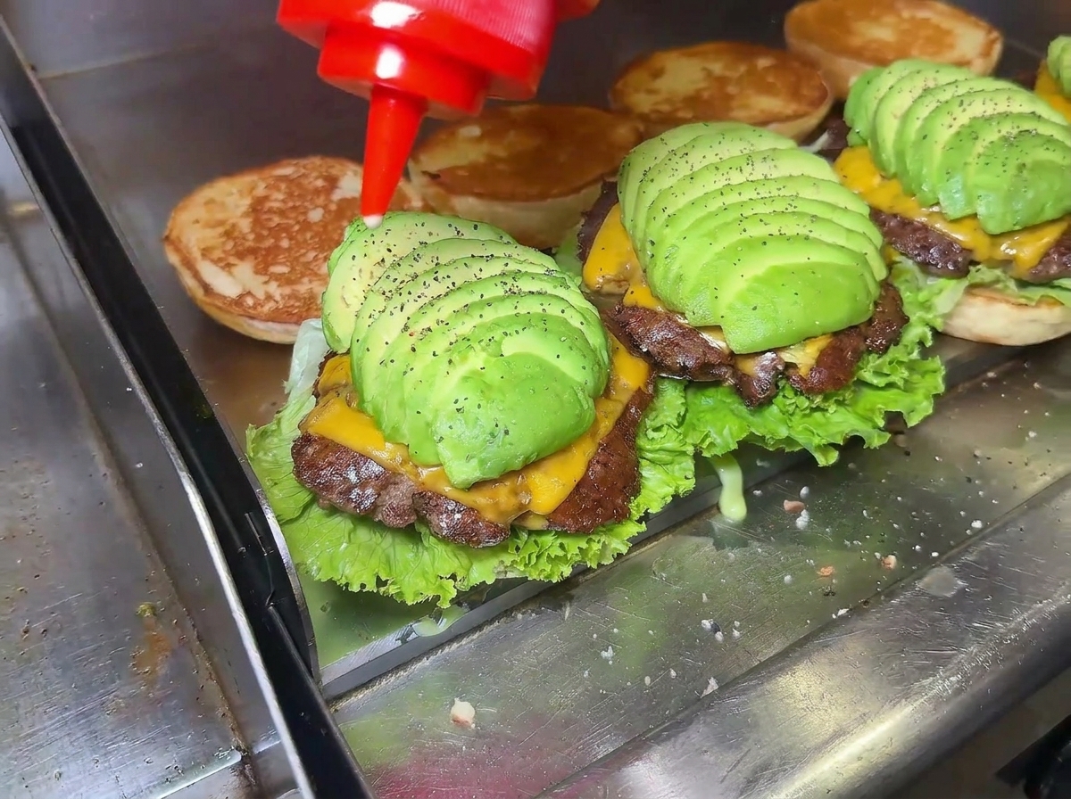 Dettaglio di una mano che versa della salsa rossa su un cheeseburger allavocado perfettamente assemblato.