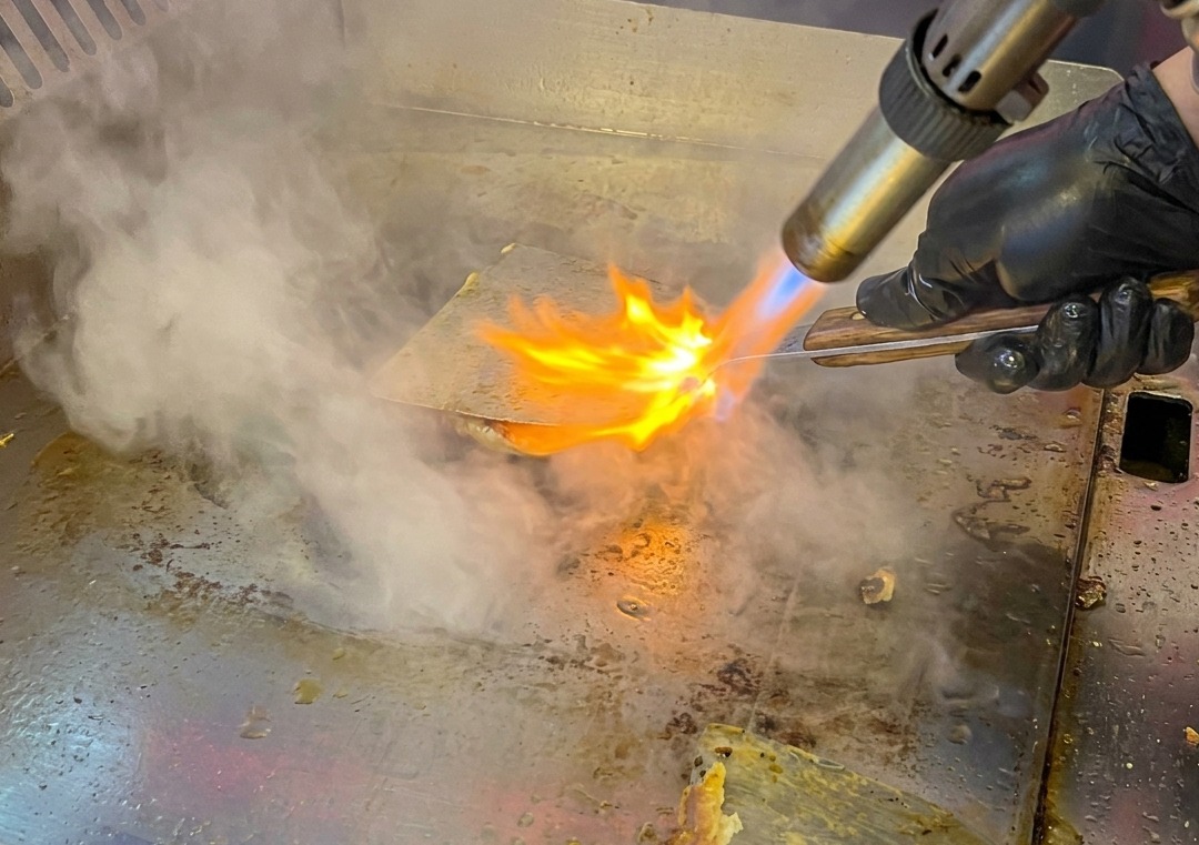 Un hamburger sulla piastra che viene fiammeggiato con un cannello, emettendo fumo e scintille.