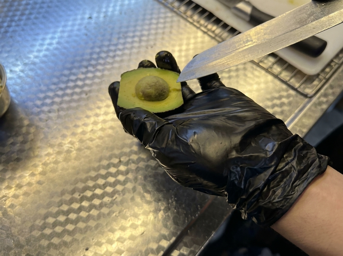 Una mano con un guanto nero tiene mezzo avocado mentre rimuove il nocciolo con un coltello.