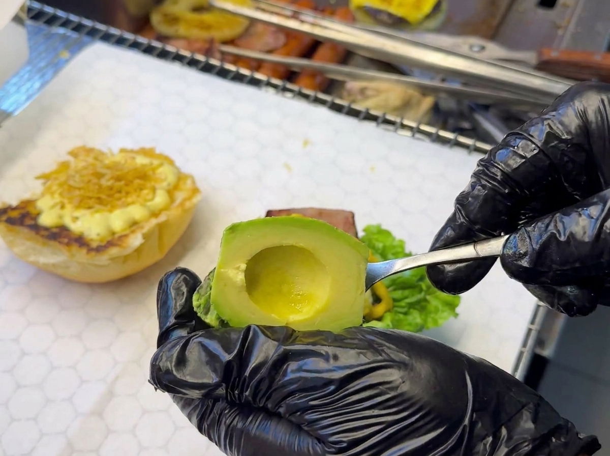 Una mano con guanto nero usa un cucchiaio per adagiare una fetta di avocado su una base per hamburger che include già lattuga e salsa.