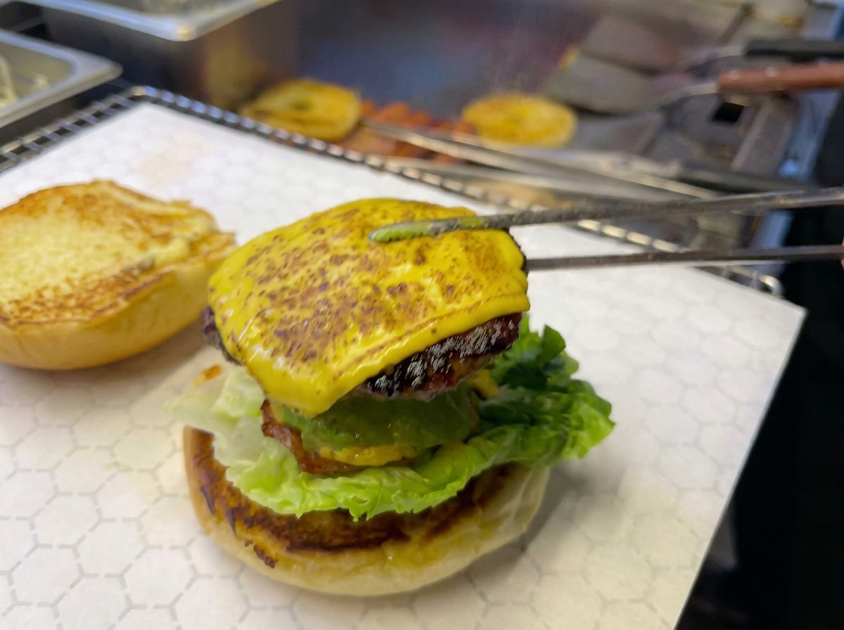 Pinze da cucina appoggiano un hamburger al formaggio sormontato da un anello di ananas su un panino con avocado e lattuga.